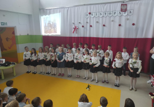 Dzieci trzymając róże w kolorze białym i czerwonym śpiewają pieśni patriotyczne.