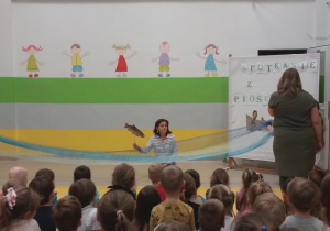Pani Anetka klęcząc na kolanach manipuluje sylwetą ryby. Dzieci trzymają niebieskie tkaniny naśladując rzekę.