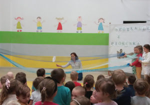 Pani Anetka klęcząc na kolanach manipuluje sylwetą ryby. Dzieci trzymają niebieskie tkaniny naśladując rzekę.