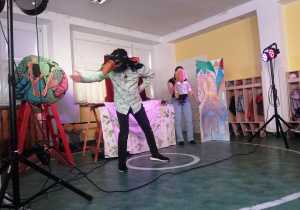 Aktor przebrany za cara kruków stoi na środku sceny. Obok aktorka trzyma kukiełkę chłopca.