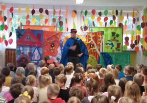 Aktor ubrany w niebieską pelerynę i czarny kapelusz rozmawia z dziećmi siedzącymi na widowni.