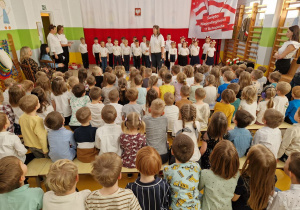 Widok z końca sali na dzieci oraz nauczycielki prowadzące uroczystość