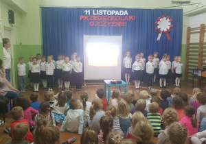 Dzieci ubrane na galowo ustawione w szachownicę recytują z wychowacą wiersz Władysława Bełzy "Katechizm polskiego dziecka".