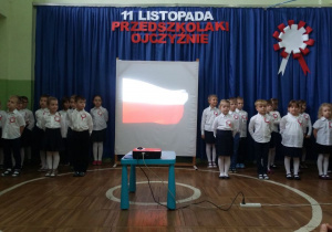 Dzieci stoją w pozycji "Na baczność", a pomiędzy nimi stoi ekran, na którym wyświetlana jest animacja poruszającej sie flagi Polski.