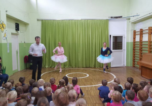Dzieci siedzą na widowni, prowadzący stoi i prezentuje stroje tancerek.