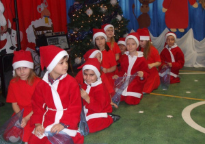 Dzieci przebrane za Mikołaje przygotowują się do występu klęcząc na podłodze.