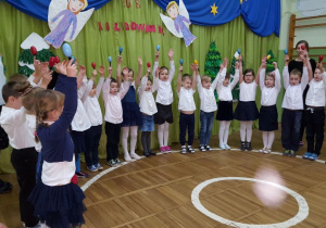 Dzieci ubrane na galowo śpiewają kolędę grając na marakasach.