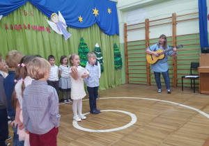 Dzieci ubrane na galowo śpiewają kolędę w asyście wychowawcy grającego na gitarze.