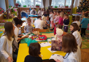 Dzieci siedzące przy stolikach obserwują przykładowe zabiegi lekarskie wykonywane na swoich pluszakach.