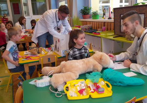 Dzieci siedzące przy stoliku obserwują badanie swojego pluszaka przez lekarza.