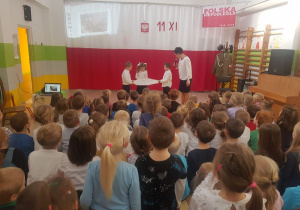 Troje dzieci na scenie razem z wychowawcą przecina karton symbolizujący rozbiór Polski.
