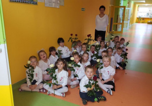 Dzieci czekające na występ z biało-czerwonymi różami w dłoniach, za którymi stoi wychowawca.