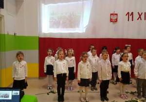 Dzieci śpiewają pieśń patriotyczną, a przed nimi leżą biało-czerwone róże.
