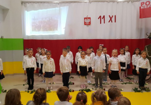 Dzieci śpiewają pieśń patriotyczną, a w tle widnieje prezentacja multimedialna przedstawiająca zdjęcia z czasów wojny.