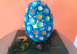 Pisanka z wykorzystaniem styropianowego jajka malowana farbą plakatową, ozdabiana niebieskimi nakrętkami od butelek plastikowych, kolorowymi kwiatkami wykonanymi przy użyciu dziurkacza ozdobnego.