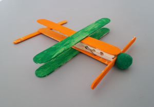 Samolot wykonany przy użyciu spinacza do bielizny, patyków po lodach, pompona, zielonej i pomarańczowej farby.