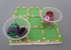 Gra wykonana przy użyciu kawałka styropianu, zielonej kartki, kolorowych dżetów, nakrętek po butelkach.